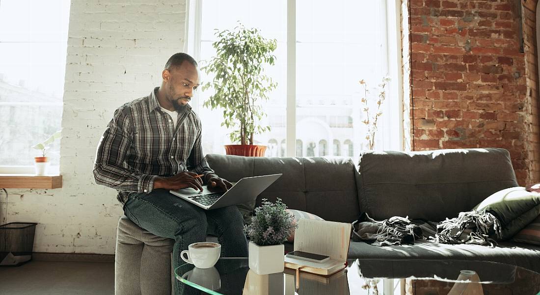 A person is sitting on a green couch working on a laptop in a brightly lit room in his home.