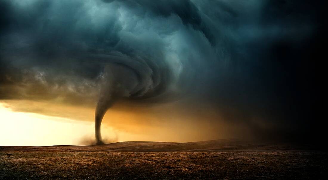 A tornado in the distance sweeping across a dry landscape. The darkening clouds brought on by the storm are mingling with an orange sunset on the horizon.