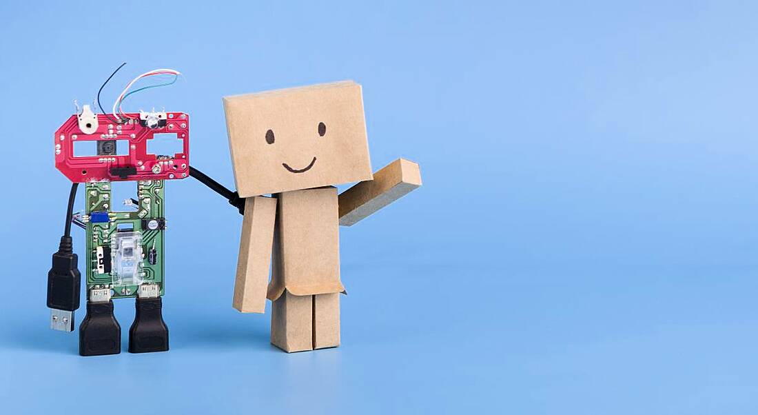 A small robot made of colourful parts is standing beside a small cardboard robot with a smiling face against a blue background.