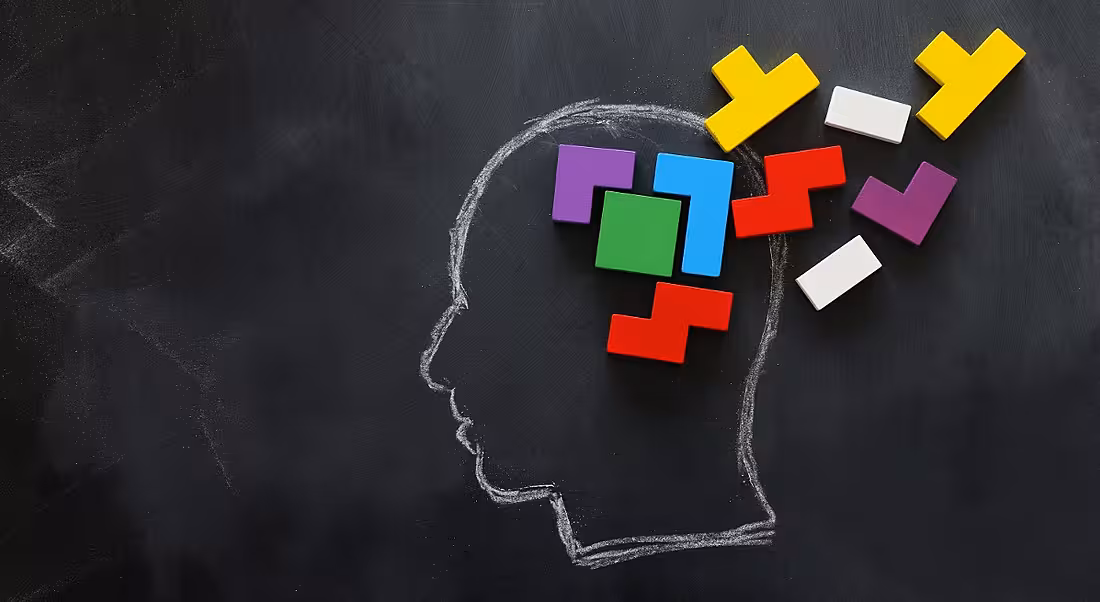 Brain made from colourful wooden puzzle inside chalk drawing of a human head on a blackboard.