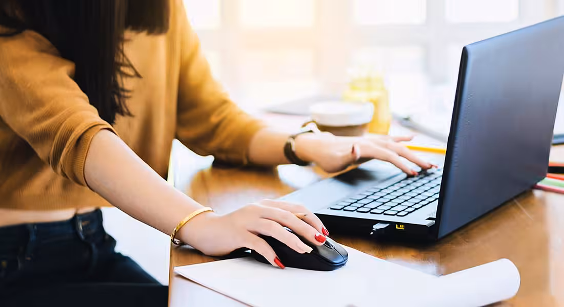 Close-up of a young woman’s hands as she uses a mouse connected to a laptop. Her fingernails are painted red and she is wearing a cropped mustard sweater and dark denim jeans.