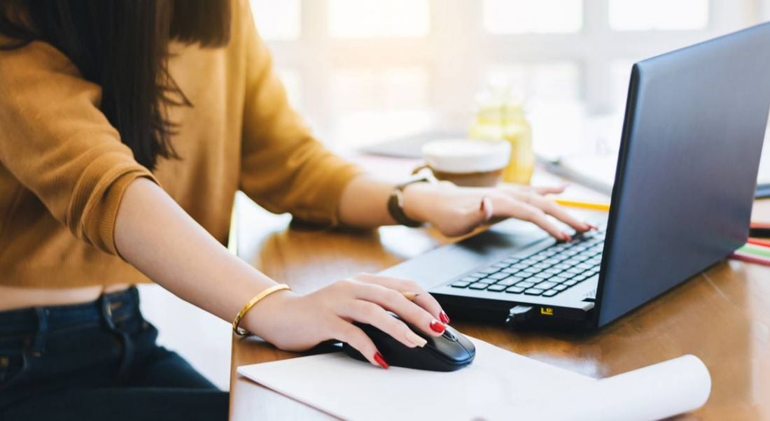 Close-up of a young woman’s hands as she uses a mouse connected to a laptop. Her fingernails are painted red and she is wearing a cropped mustard sweater and dark denim jeans.