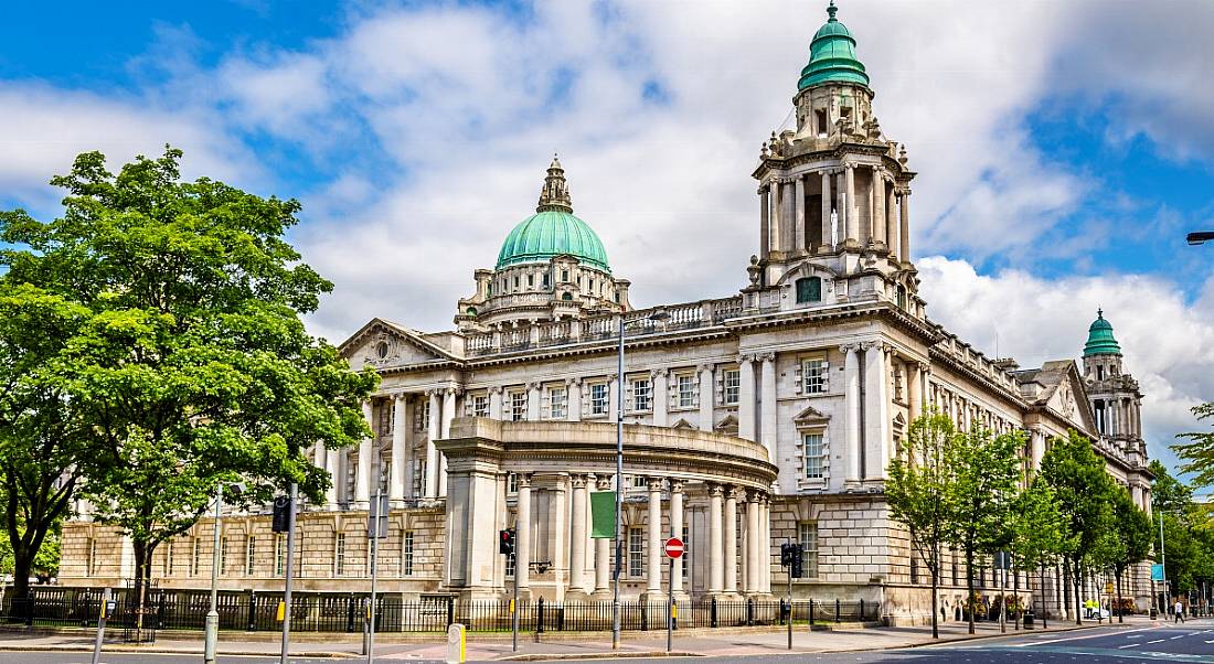 Belfast City Hall shot on a bright day, with a blue sky. There are leaves on the surrounding trees and the roads are empty.