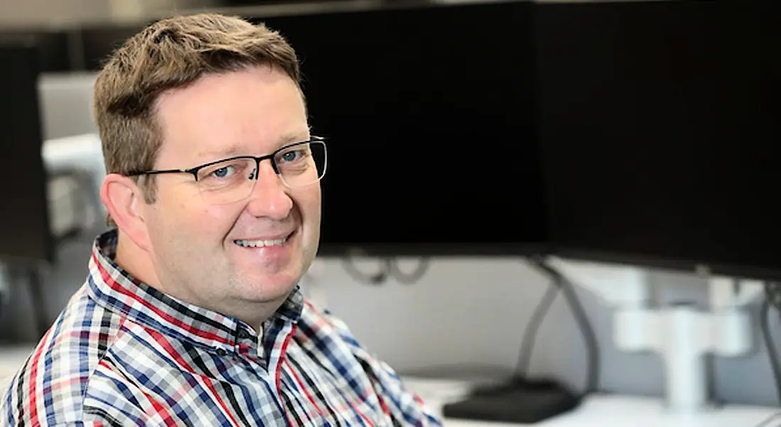 Chris Cassidy of Takeda Dunboyne is sitting in front of a computer on a desk and smiling into the camera.