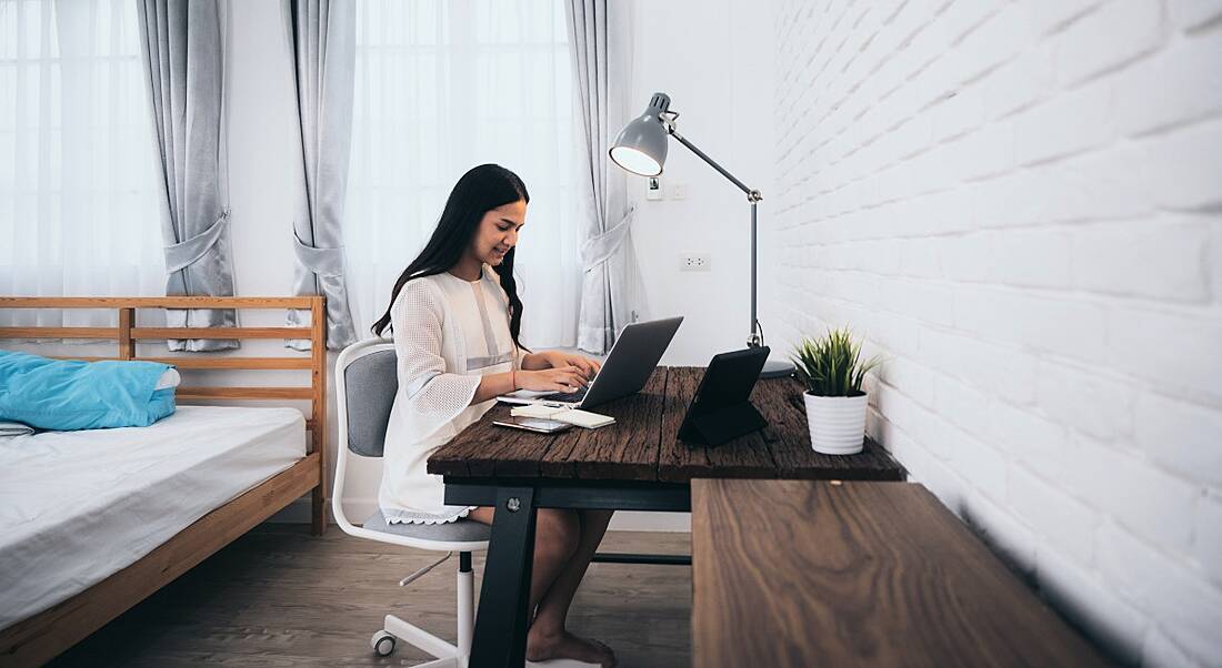 A dark-haired young woman remote working on a laptop at a desk in her bedroom.