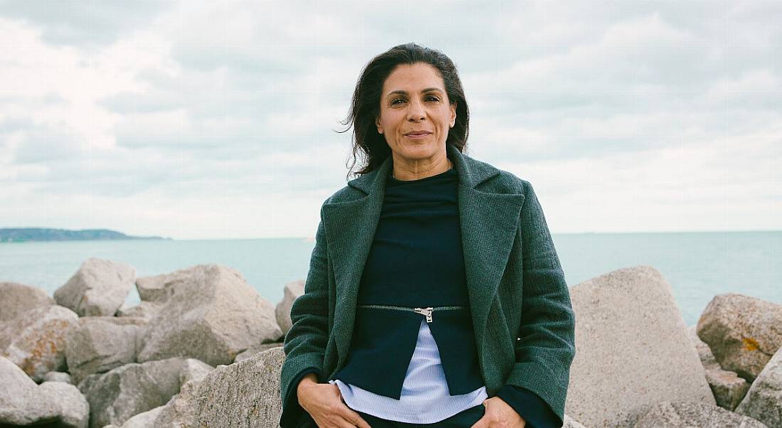 A woman with dark hair stands in front of a group of rocks in front of the ocean. She’s wearing a green coat.