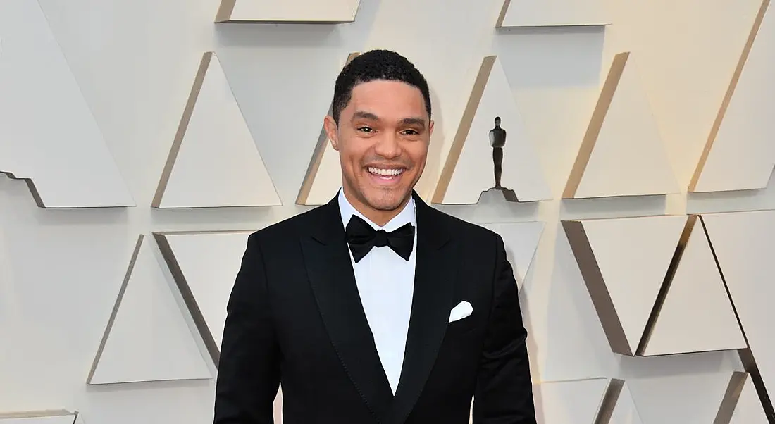 Trevor Noah stands smiling in a tuxedo against a white backdrop with geometric patterns.