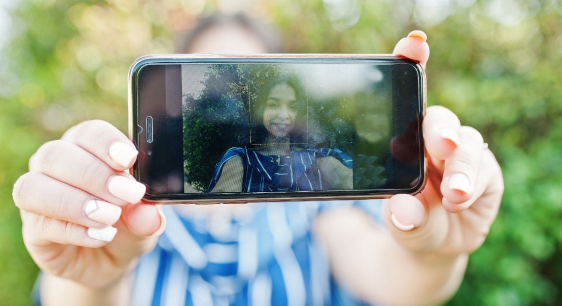 A woman standing outside in the sun, holding a smartphone in front of her face, taking a selfie.