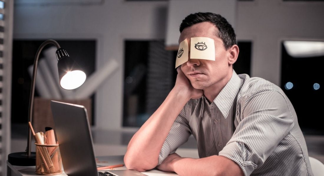 A man is sitting at his office desk with post-it notes stuck over his eyes. On the post-its are drawings of eyes to make it seem like he is awake.