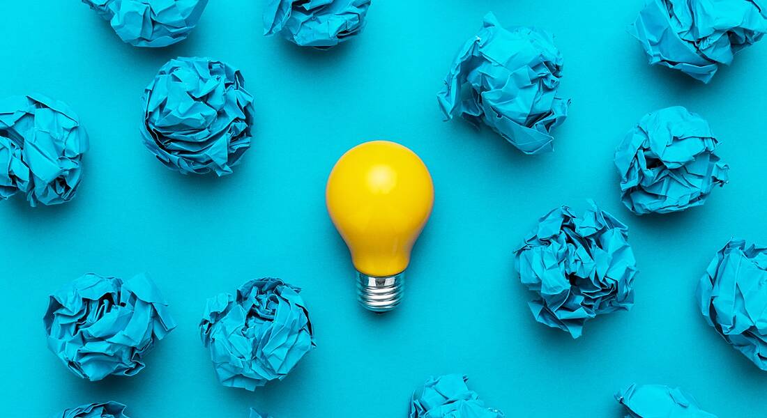 A yellow lightbulb surrounded by crumpled up blue pieces of paper against a blue background.