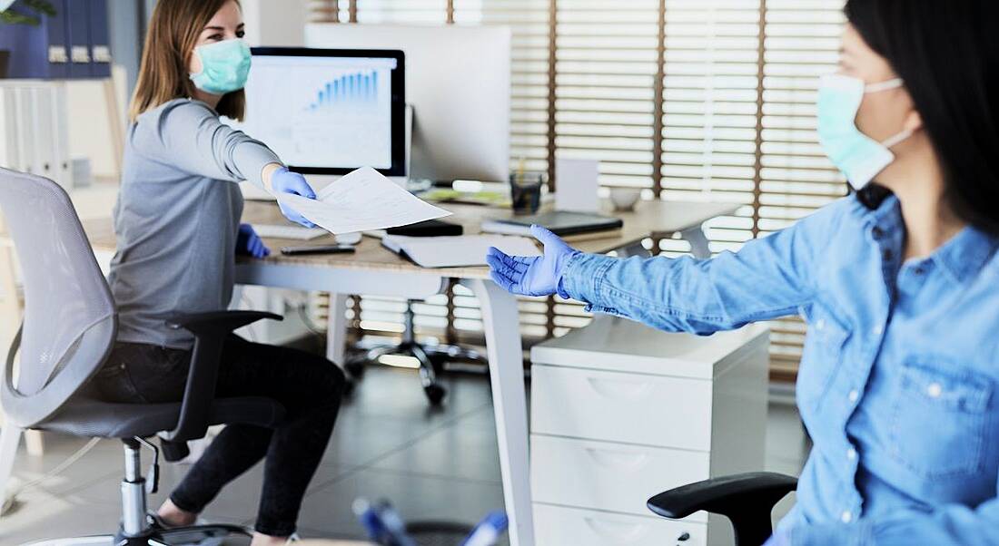 Two people in an office passing documents while wearing face masks and keeping a distance from each other.