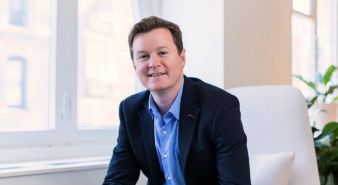 Colm O’Cuinneain, a fair-haired man in a shirt and blazer, sits on a light grey chair in a bright office space with a plant behind him.