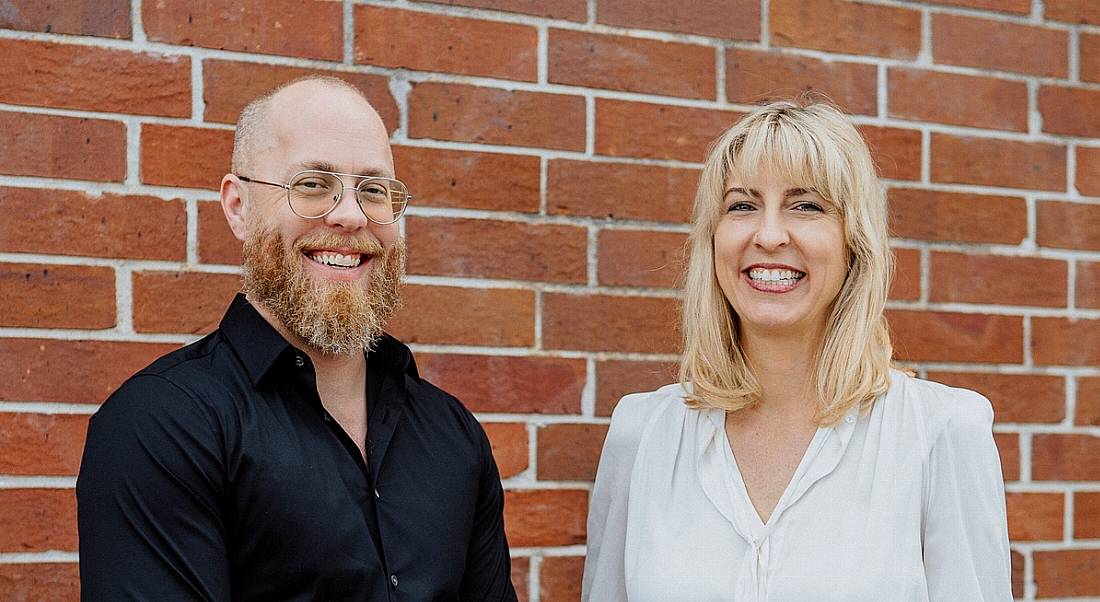 Douglas Ferguson and Karen Holst are standing against a red-brick wall and smiling into the camera.