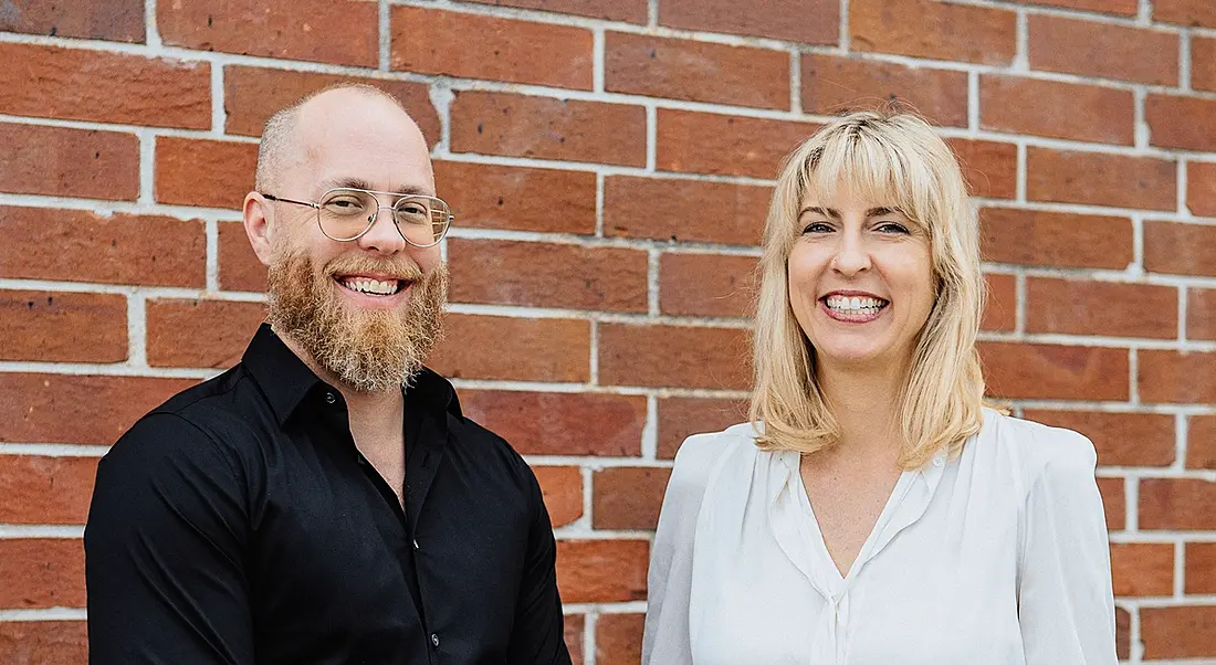 Douglas Ferguson and Karen Holst are standing against a red-brick wall and smiling into the camera.