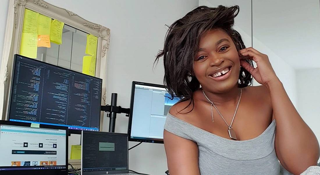 DevOps engineer Jennifer Opal is smiling into the camera beside her work desk with various computer screens in an office.