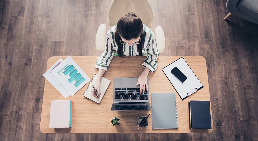 A view from a high angle of a woman remote working at a desk at home with a laptop and an assortment of files.