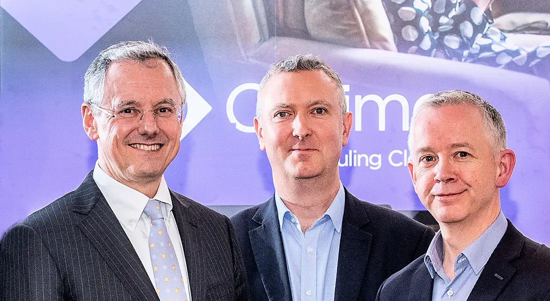 Three men wearing navy suits and white shirts standing in front of a lilac sign. The three men have short grey haircuts.