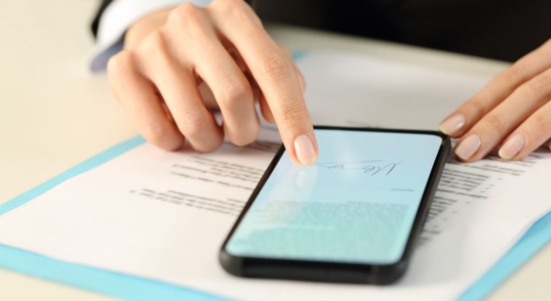 A mobile phone sits on top of a document as a woman uses her finger to sign a document on her phone.