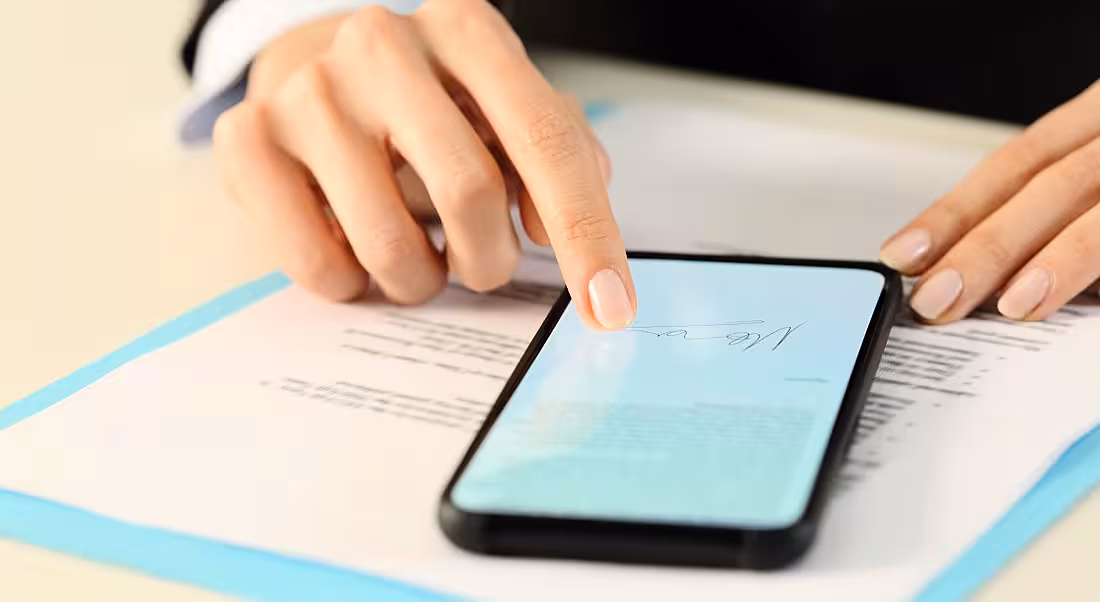 A mobile phone sits on top of a document as a woman uses her finger to sign a document on her phone.