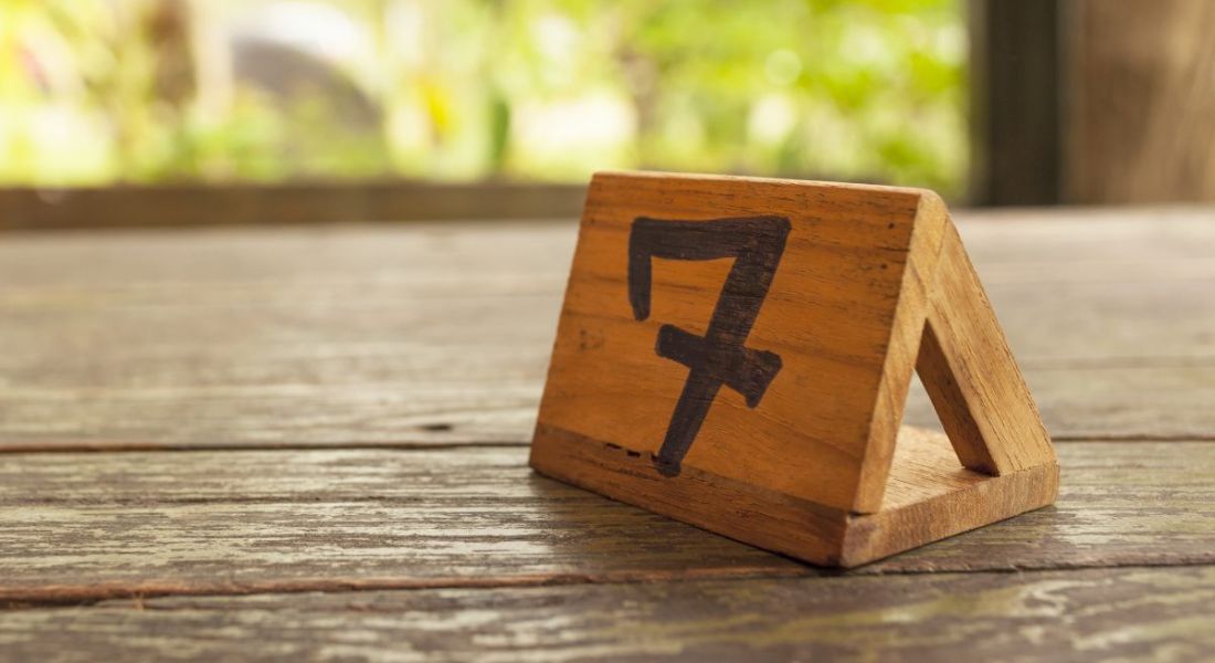 A wooden table sign with the number seven on it sits on a wooden table against a backdrop of greenery.