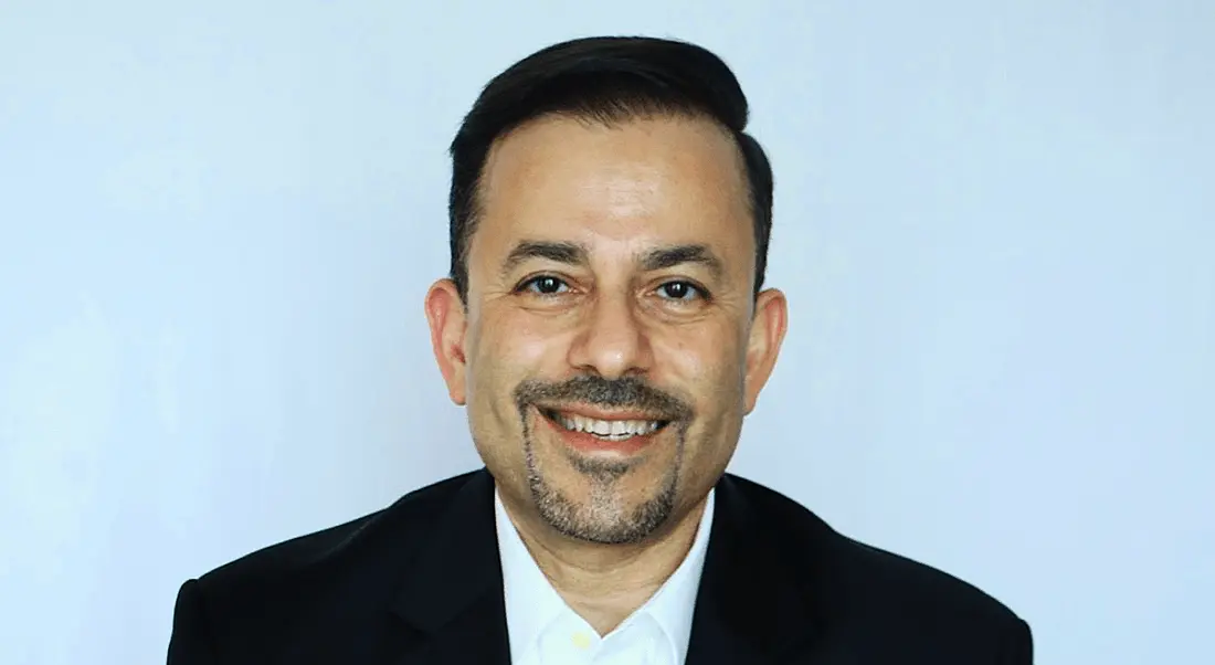 A man with black hair and stubble smiling into the camera. He is wearing a black suit and white shirt.