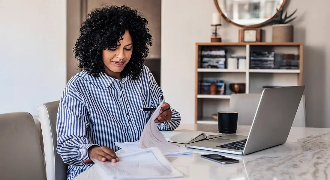 A woman sitting at a desk at home going through documents and working at a laptop.