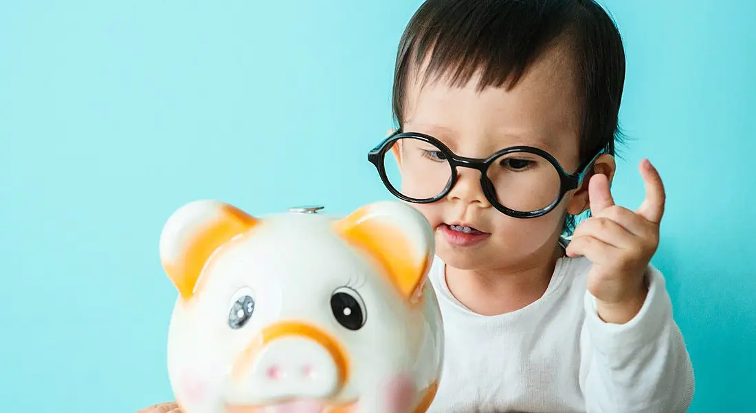 A baby wearing glasses putting a coin in a white and orange piggy bank.