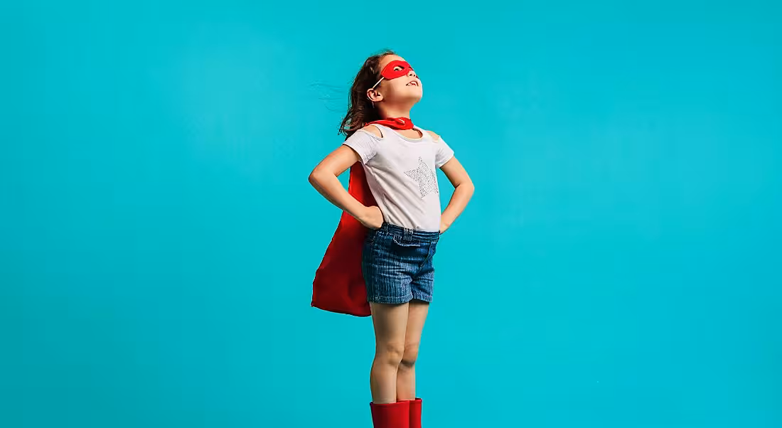 A young girl is dressed up as a superhero against a blue background, symbolising self-confidence.