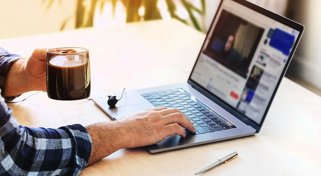 A person is taking part in online training or education at home on their laptop, with a cup of coffee in their hand.