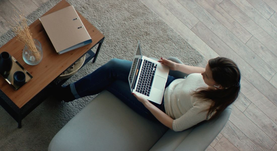 A woman is working remotely in her home on her laptop while sitting on a grey couch beside a wooden table.