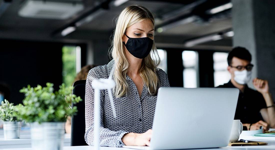 Woman wearing a face mask in an office at a laptop at a distance from someone else wearing a face mask.