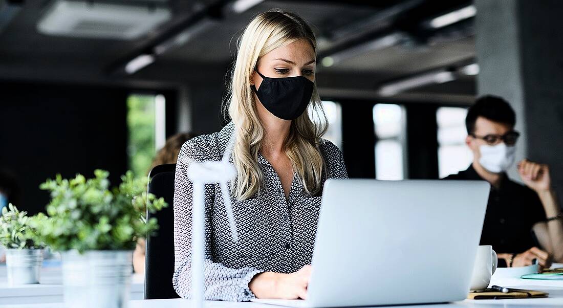 Woman wearing a face mask in an office at a laptop at a distance from someone else wearing a face mask.