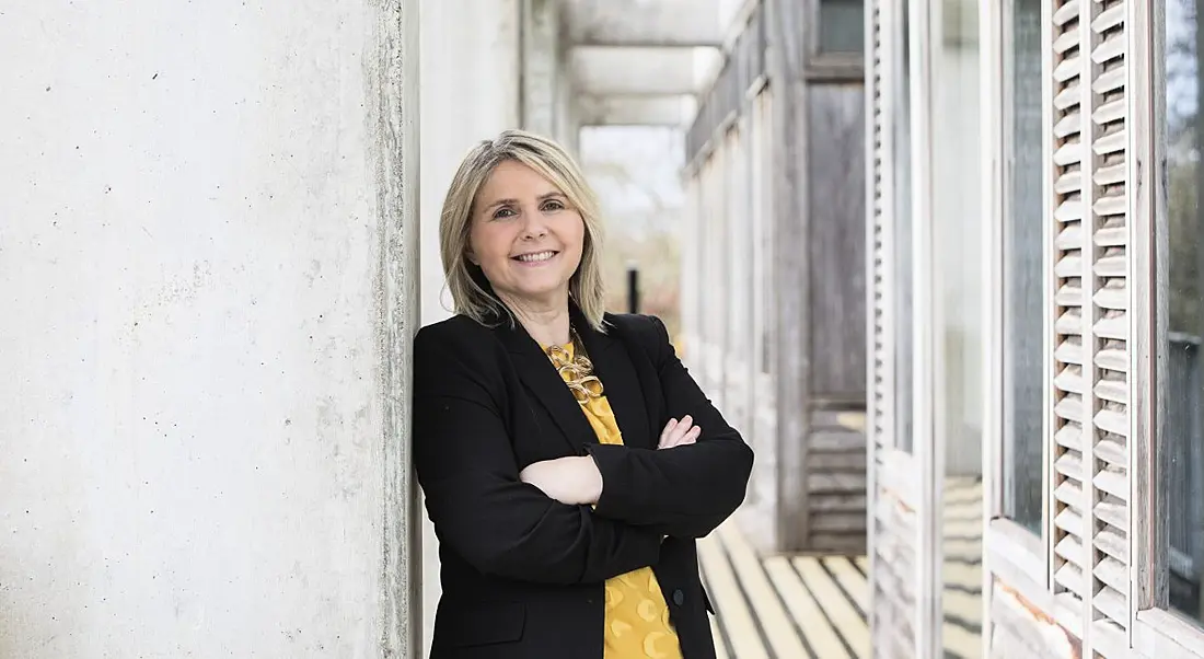 Prof Sarah Culloty is standing against a wall in UCC with her arms folded while smiling into the camera.