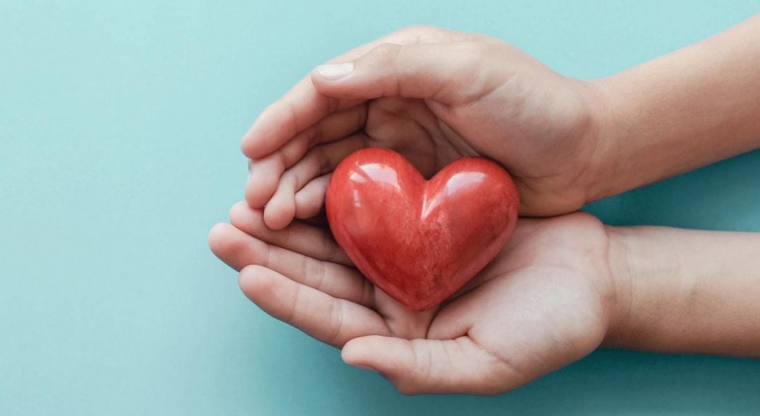 A pair of hands holding a red, wooden heart against a bright blue background.