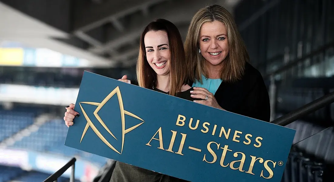 Fiona Descoteaux and Annabelle Conway stand in stadium stalls holding a banner that says Business All-Stars.