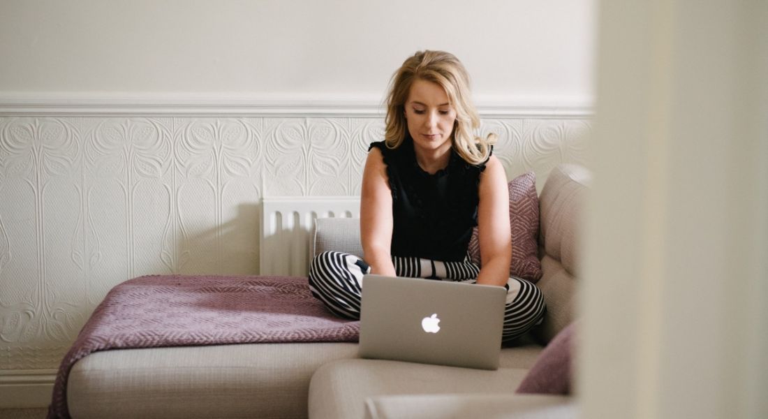 A blonde-haired woman in a black top and patterned trousers sits cross-legged on a beige L-shaped couch, working on a Macbook.