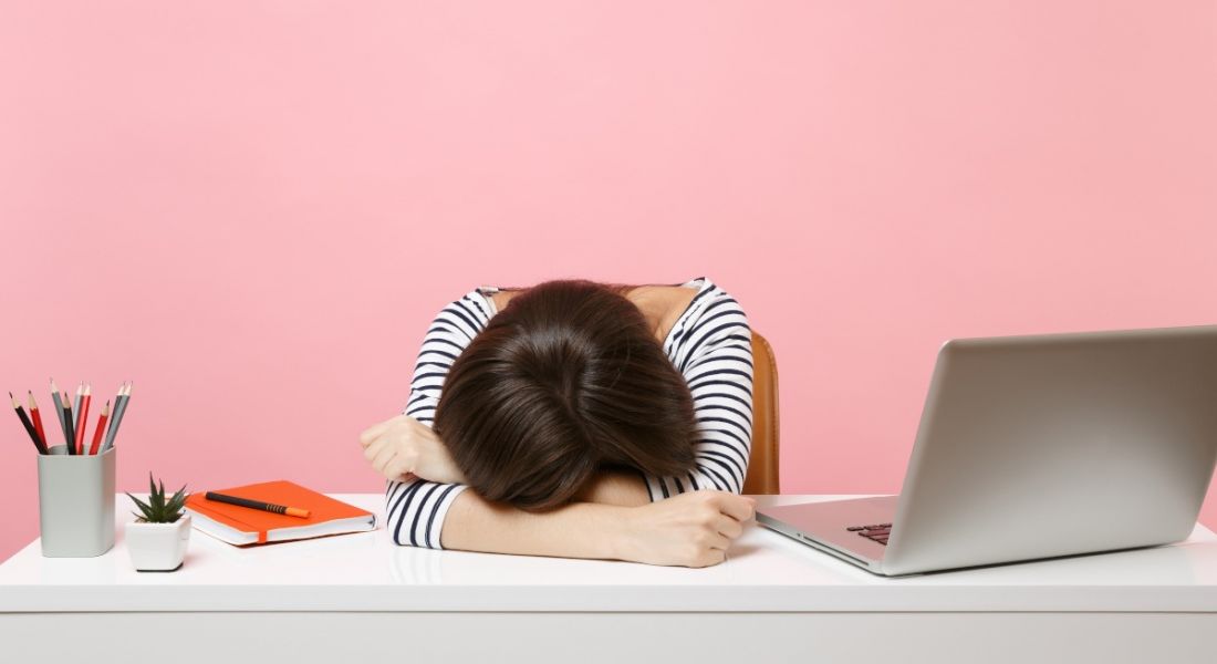 A young woman with dark hair is laying her head on a desk beside her laptop. She is sitting in a bright pink office space.