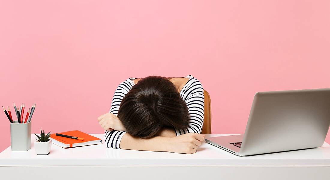 A young woman with dark hair is laying her head on a desk beside her laptop. She is sitting in a bright pink office space.