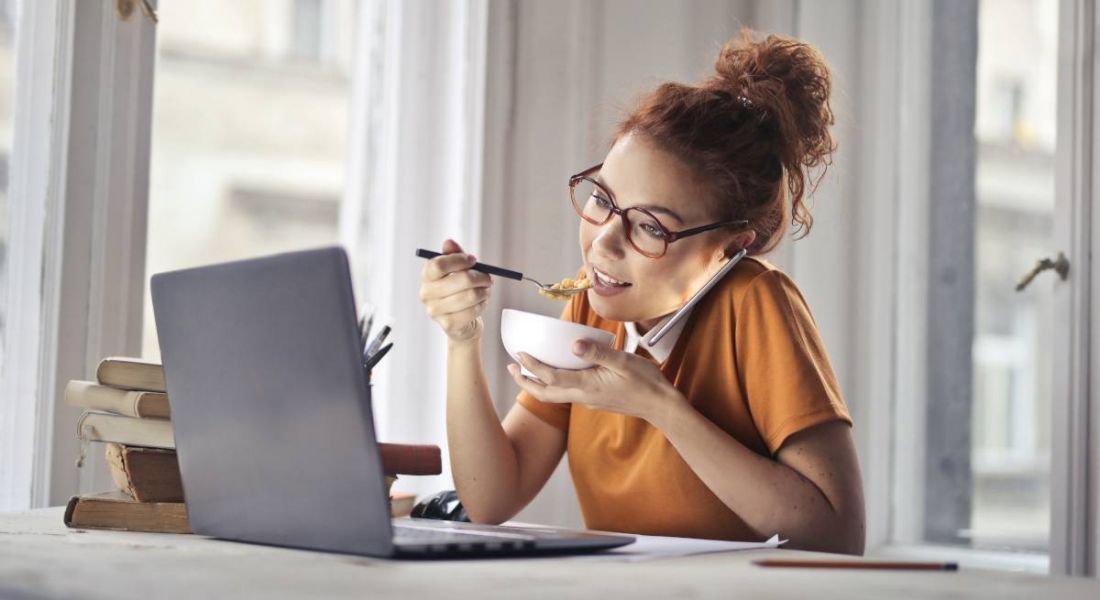 A woman in casual clothes is sitting at a desk in a bright room at home. She is eating lunch in front of her laptop while talking on the phone.