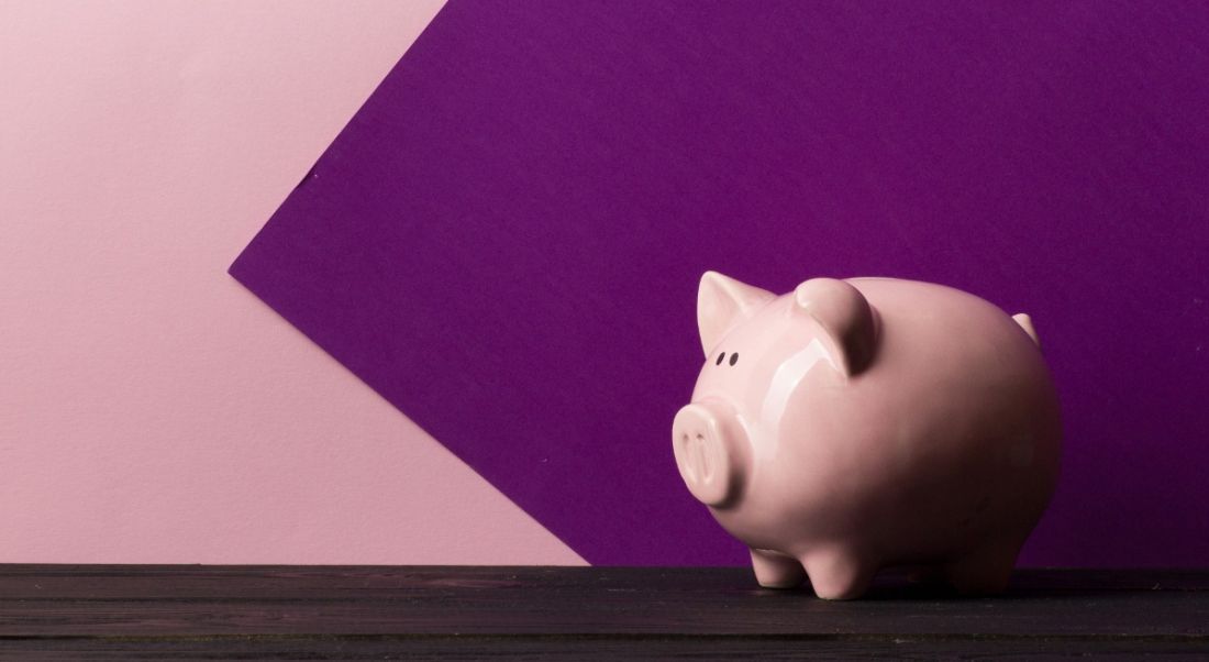 A piggy bank is standing on a dark wooden table against a pink and purple background.