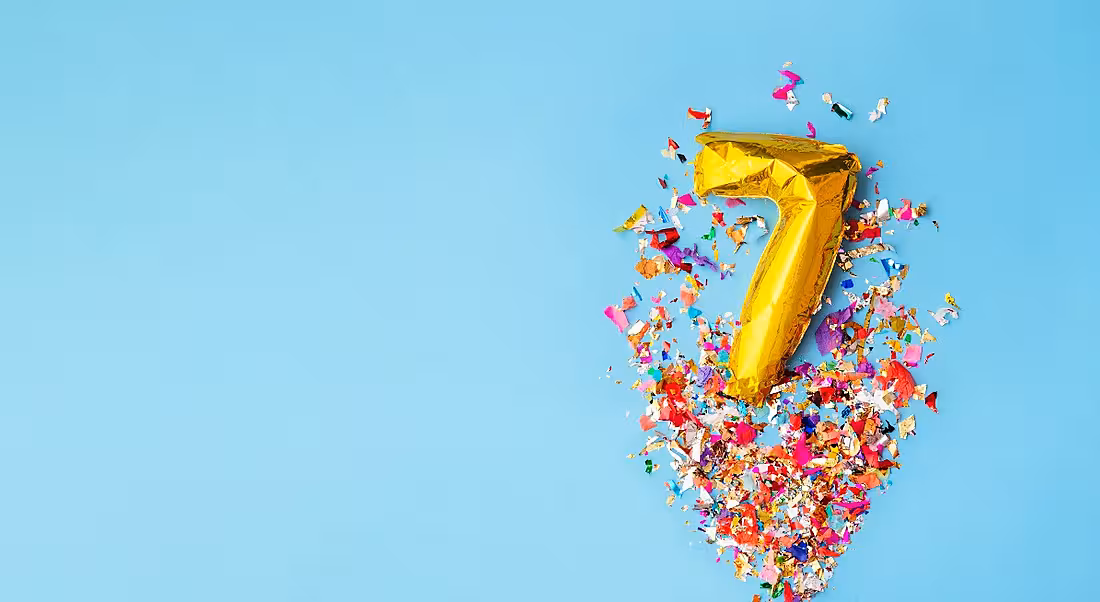 Number seven gold foil balloon with confetti on a blue background.