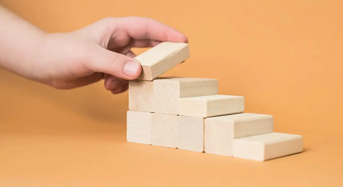 Hand arranging wooden blocks into a stepped shape against a pale orange background.