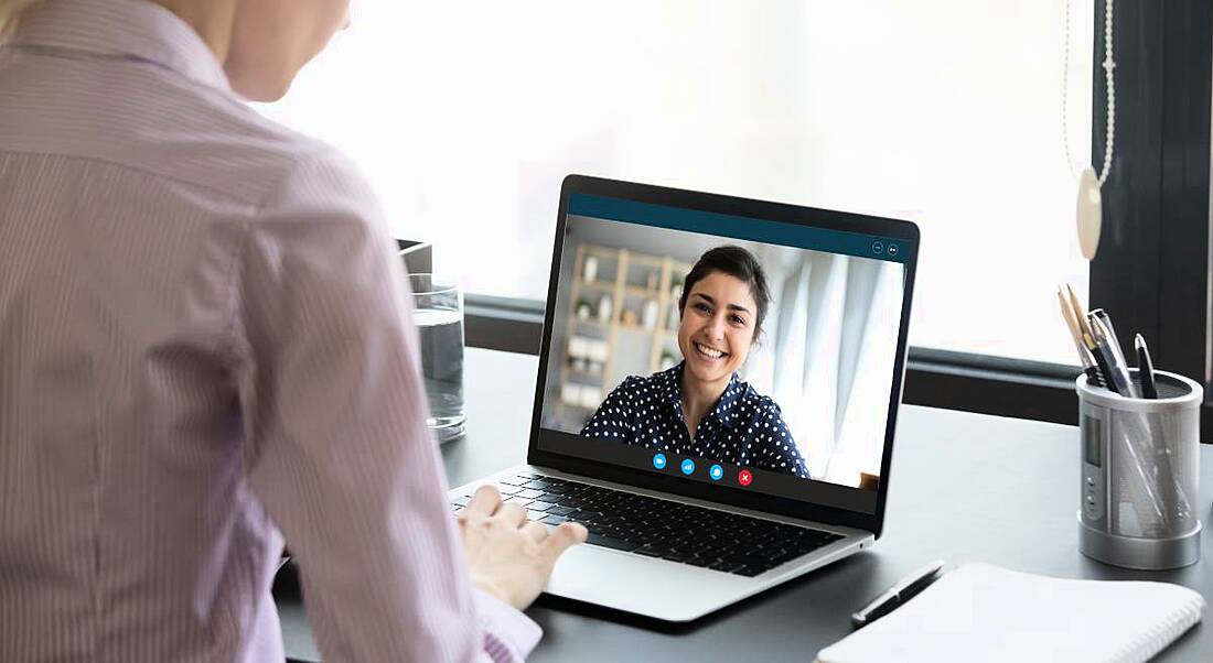 A woman in a shirt sits at a desk in front of her laptop. On the laptop screen is a video-call app, showing a second woman who she is talking to.