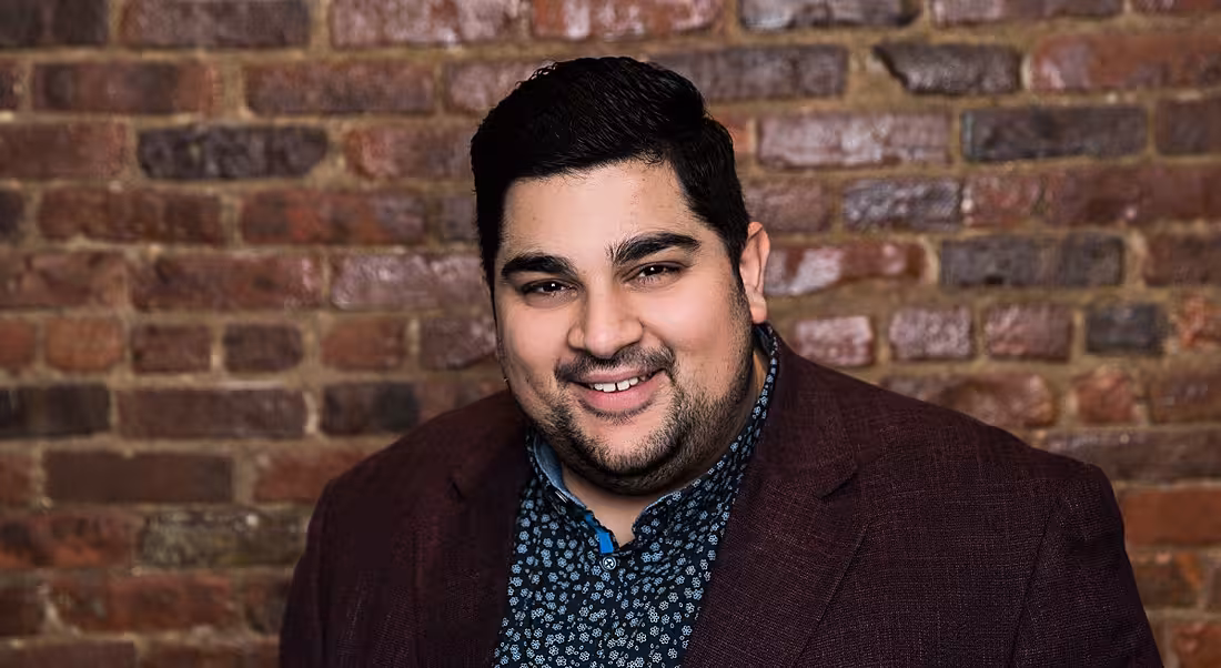 Ali Fazal of Hibob is standing against a red-brick wall and smiling into the camera.