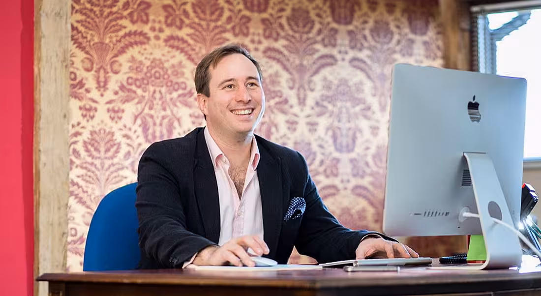 Patrick Tame is sitting at a computer in an office with patterned wallpaper in the background.