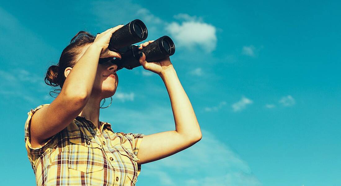 A woman in a check shirt looking through binoculars against a blue sky with fluffy white clouds.