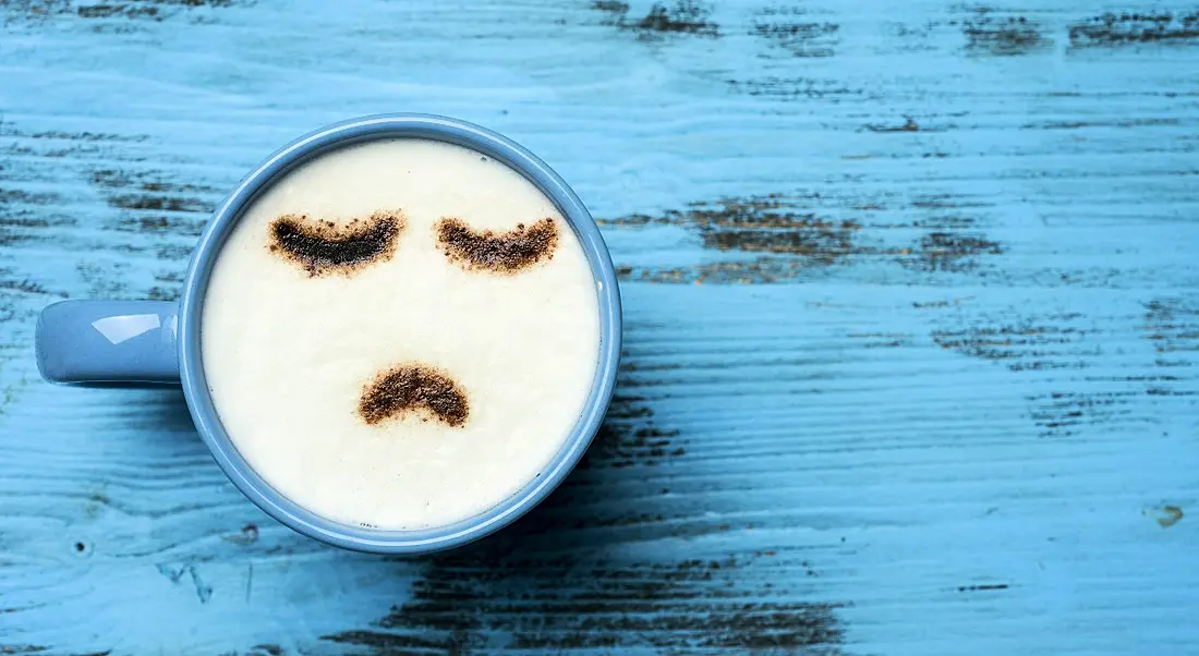 Blue cup of cappuccino with a sad face drawn with cocoa powder on its milk foam, on a blue rustic table.