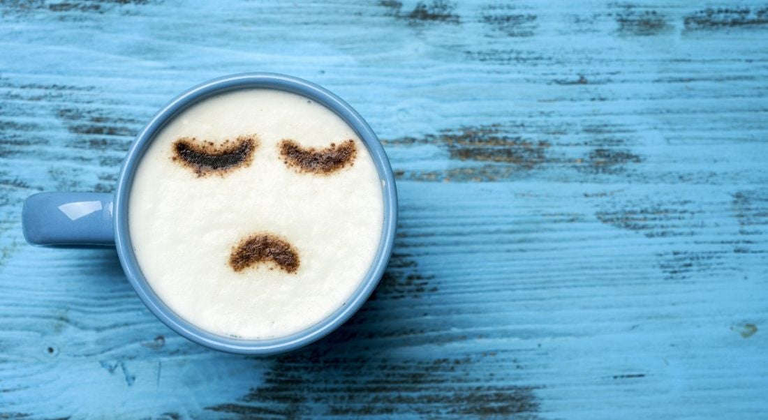 Blue cup of cappuccino with a sad face drawn with cocoa powder on its milk foam, on a blue rustic table.