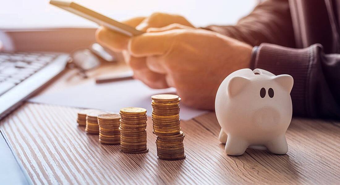 A person is doing calculations and writing notes at home beside stacks of coins and a piggy bank.