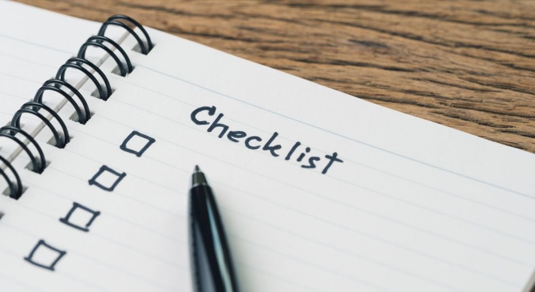 Close-up of a notebook page with a checklist written on it and a black pen on a wooden table.